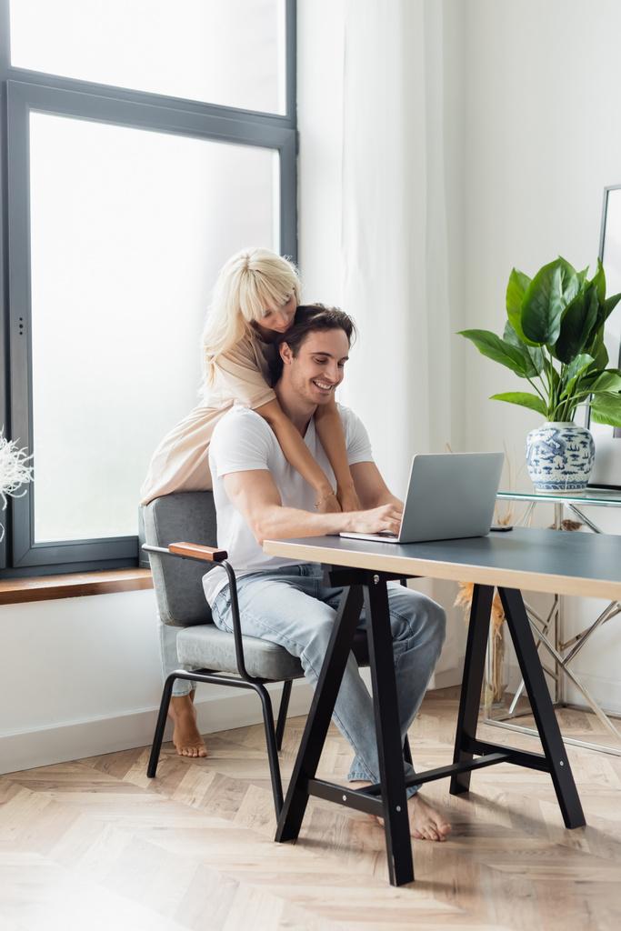 femme blonde étreignant petit ami heureux en utilisant un ordinateur portable tout en travaillant à la maison - Photo, image