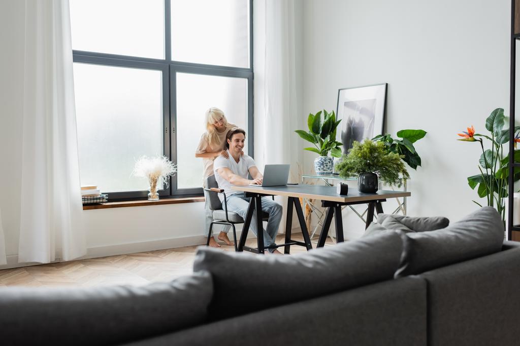 femme blonde massant les épaules de petit ami travaillant à la maison avec un ordinateur portable - Photo, image