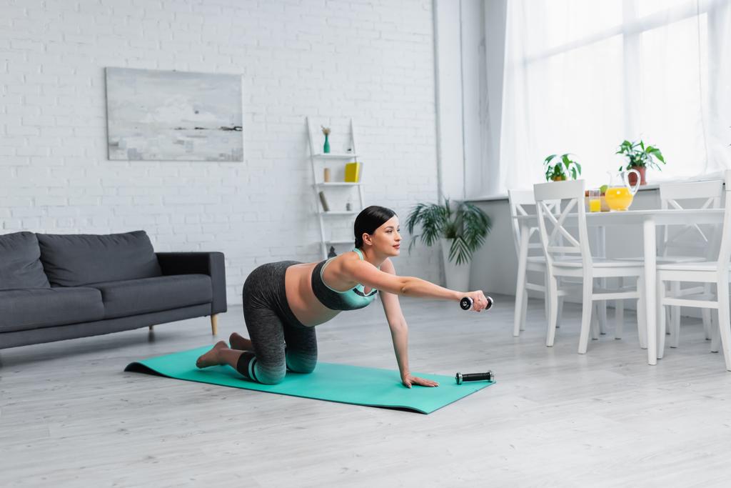 junge schwangere Frau trainiert mit Hantel, während sie auf Knien auf der Fitnessmatte steht - Foto, Bild