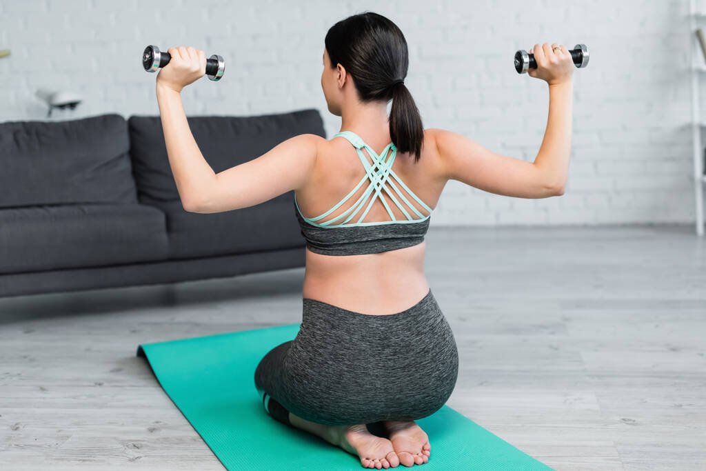 vista trasera de la mujer en ropa deportiva haciendo ejercicio con pesas mientras está sentado en la alfombra de fitness - Foto, imagen