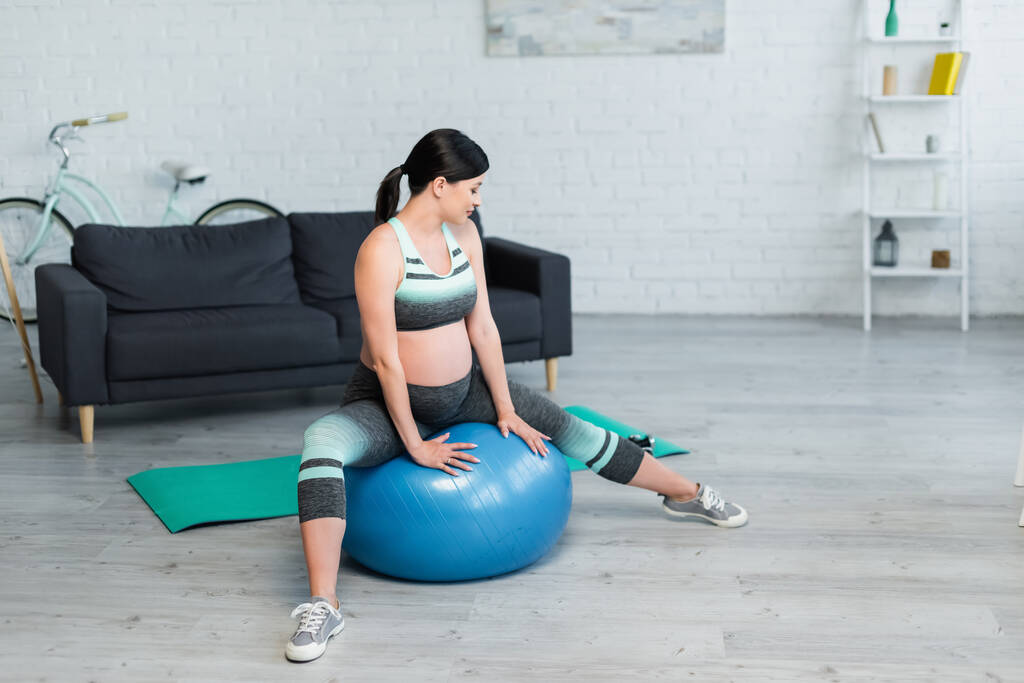 Brünette schwangere Frau in Sportbekleidung trainiert auf Fitnessball im Wohnzimmer - Foto, Bild