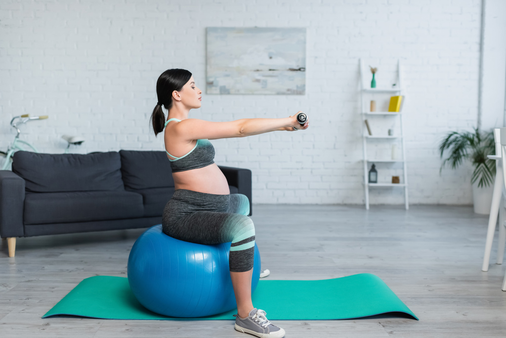 πλαϊνή όψη της μελαχρινής εγκύου γυναίκα άσκηση με αλτήρες, ενώ κάθεται σε μπάλα φυσικής κατάστασης - Φωτογραφία, εικόνα