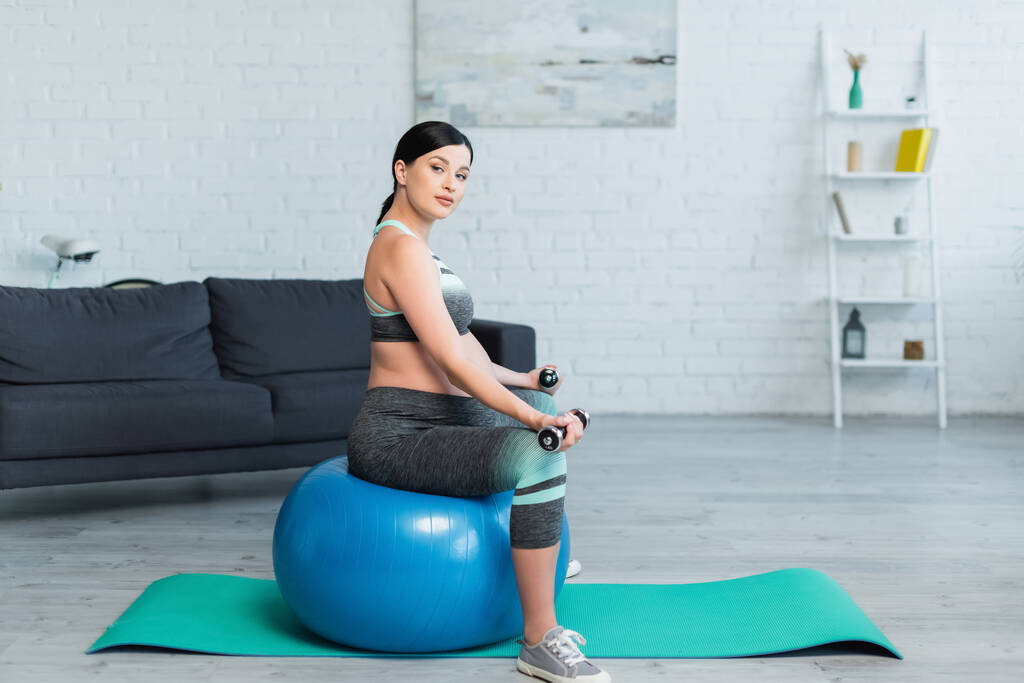 hezká těhotná žena dívá na kameru, zatímco sedí na fitness míč doma - Fotografie, Obrázek