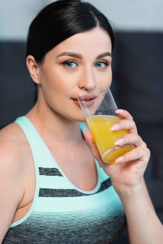 mladá žena ve sportovní podprsenka při pohledu na kameru při pití čerstvé pomerančové šťávy - Fotografie, Obrázek