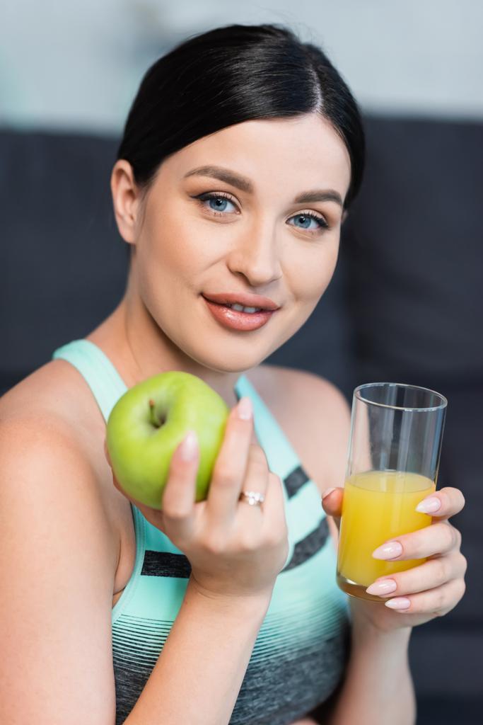 pretty, brunette woman holding ripe apple and fresh orange juice while smiling at camera - Photo, Image