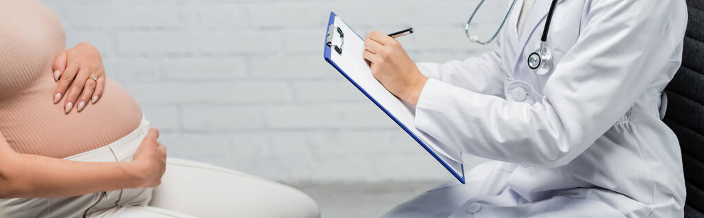 妊娠中の女性バナーの近くにクリップボードを書いている医師の部分的なビュー - 写真・画像