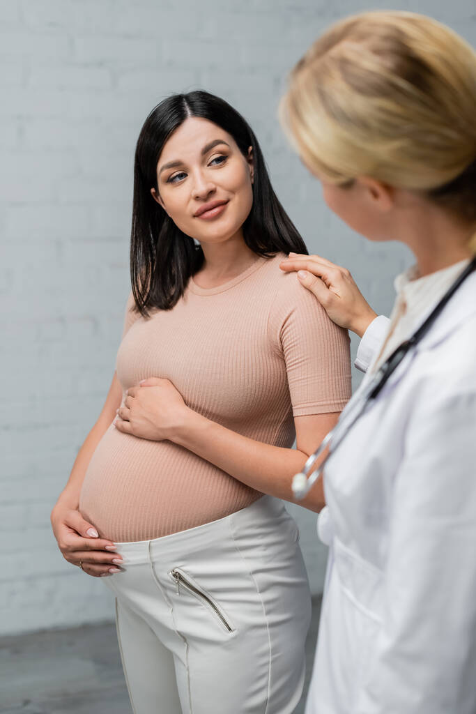 беременная женщина улыбается, когда размытый доктор трогает ее плечо во время консультации - Фото, изображение