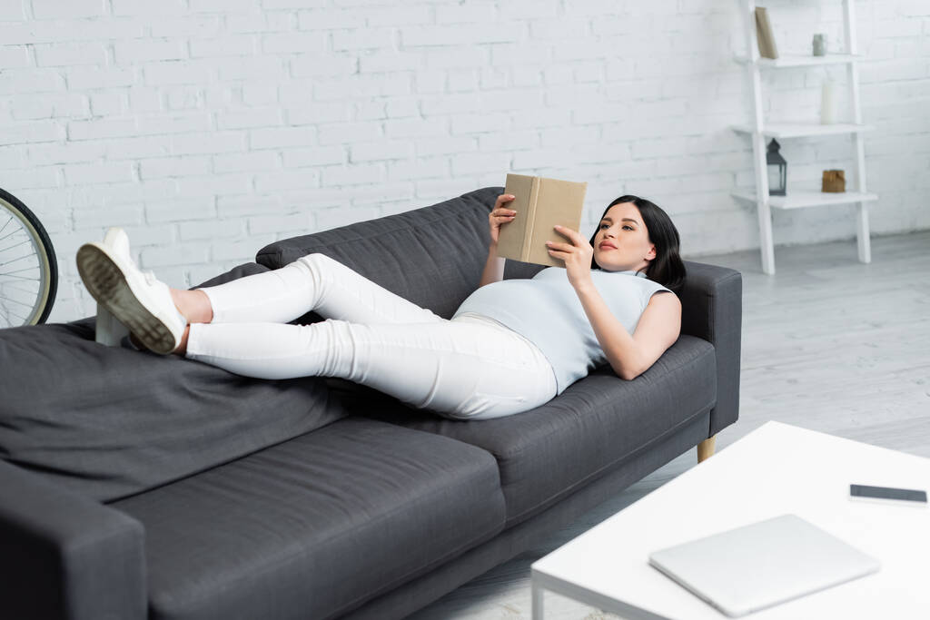 mulher grávida lendo livro enquanto deitado no sofá perto de dispositivos borrados na mesa na sala de estar - Foto, Imagem