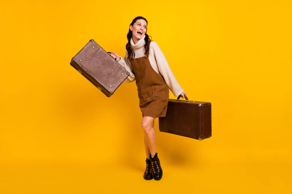 Volledige lengte foto van schattige zoete dame gekleed bruin overall holding twee koffers op zoek lege ruimte geïsoleerde gele kleur achtergrond - Foto, afbeelding
