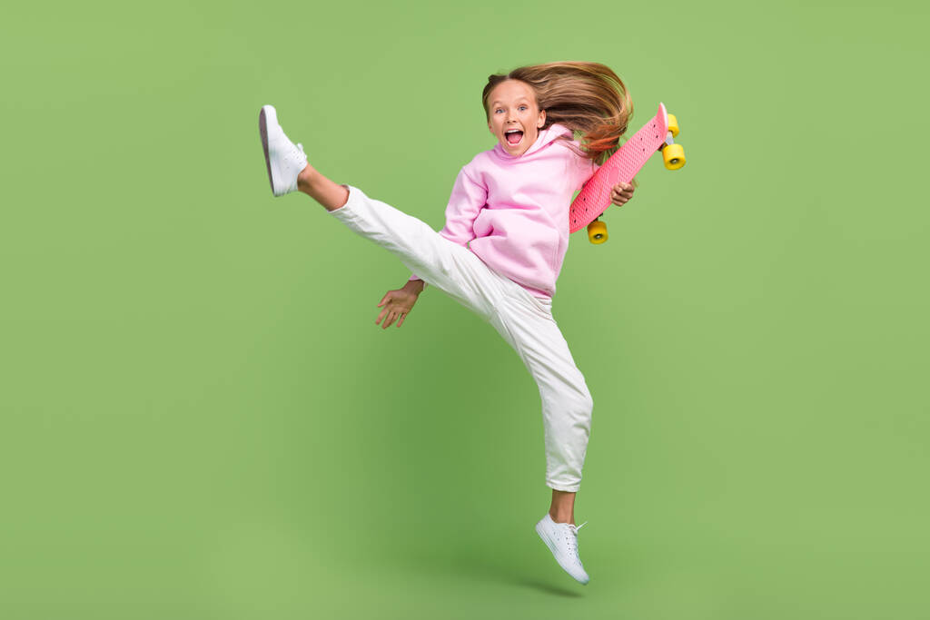 Foto de corpo inteiro de cool pequena menina loira salto com skate desgaste com capuz calças sapatilhas isoladas no fundo verde - Foto, Imagem