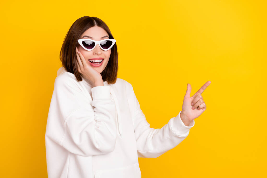 Photo of excited promoter lady indicate finger empty space wear sunglass white hoodie isolated yellow color background - Photo, Image