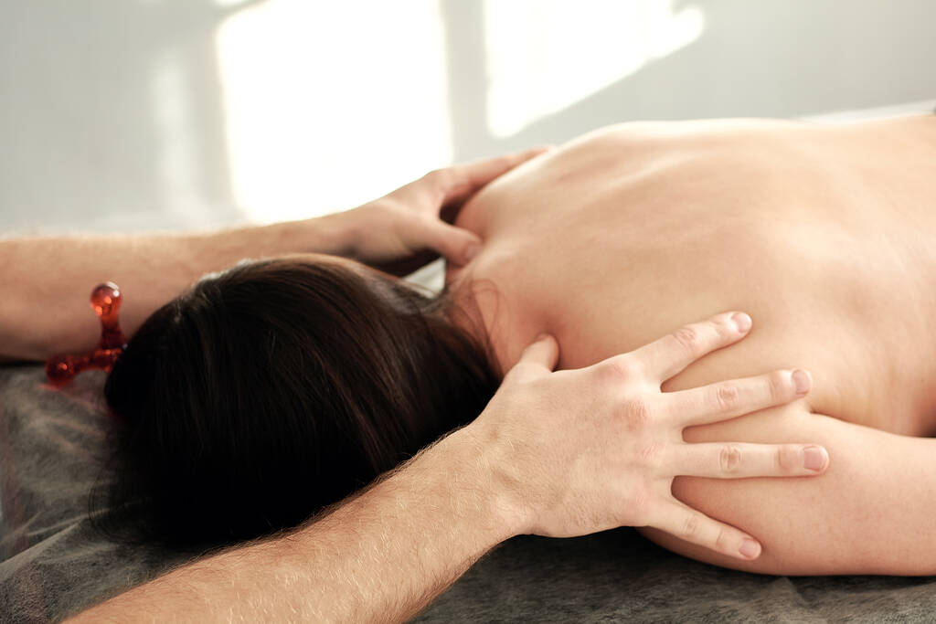 Un masajista masculino lleva a cabo tratamientos de spa para una joven que está acostada boca abajo en un sofá. - Foto, imagen
