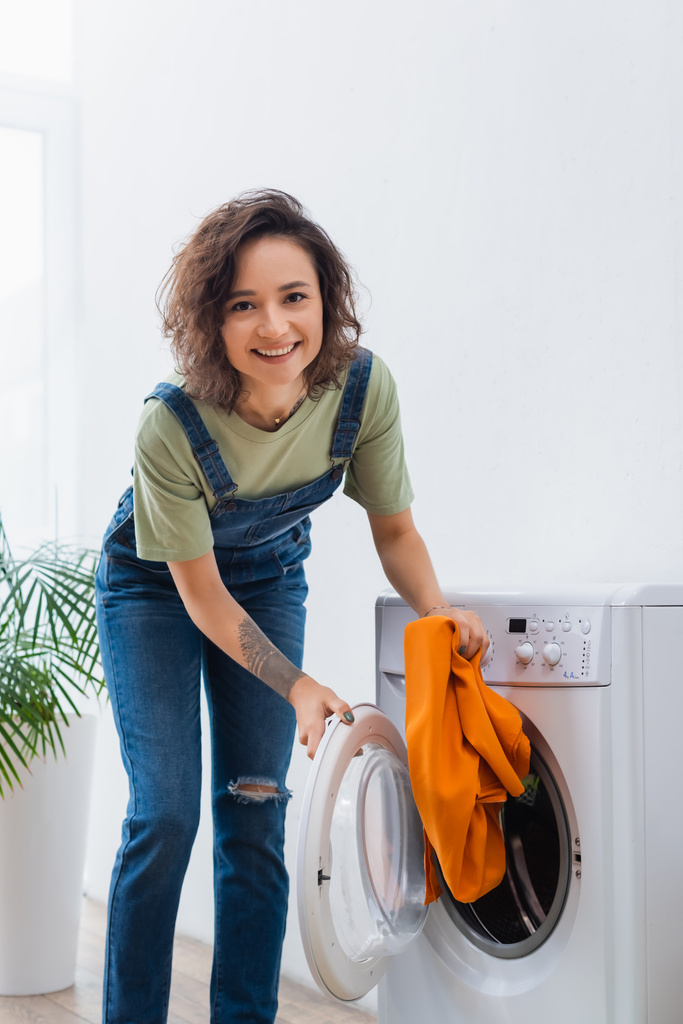 Neşeli ev hanımı çamaşır makinesinin yanında giysileri tutarken kameraya bakıyor. - Fotoğraf, Görsel