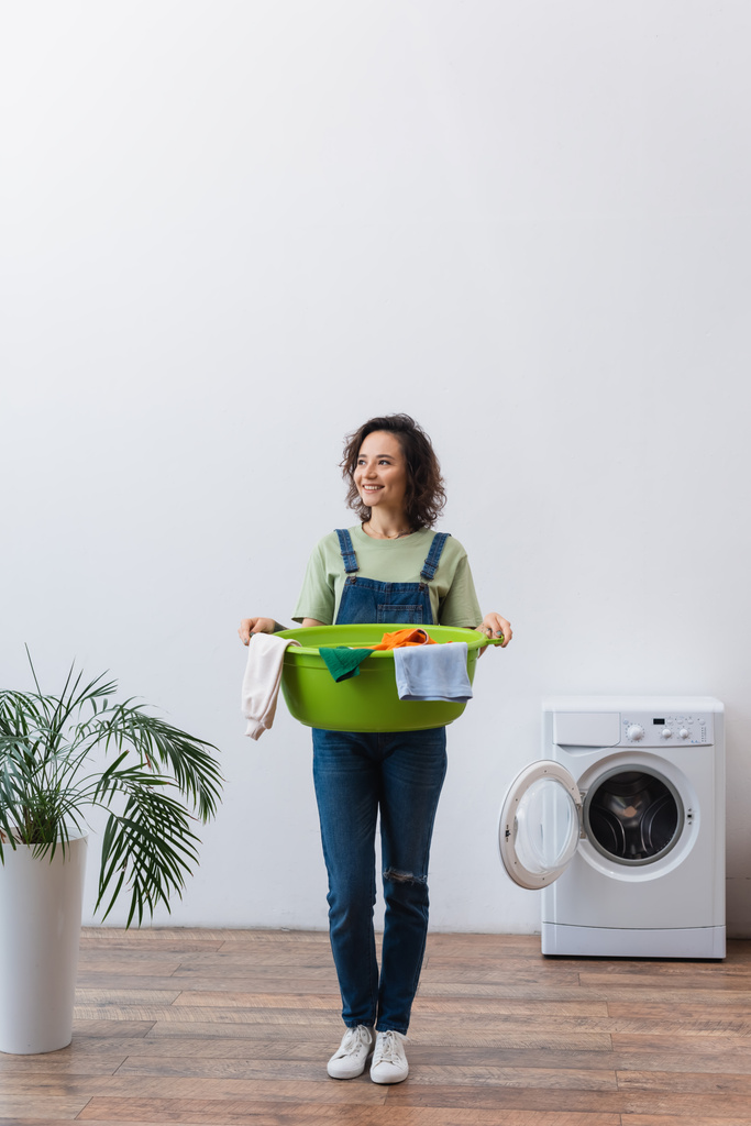 visão comprimento total da dona de casa feliz segurando bacia de lavanderia com roupas perto de planta e máquina de lavar roupa - Foto, Imagem