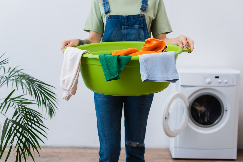 обрезанный вид на домохозяйку, держащую миску для стирки возле размытой стиральной машины в ванной комнате - Фото, изображение