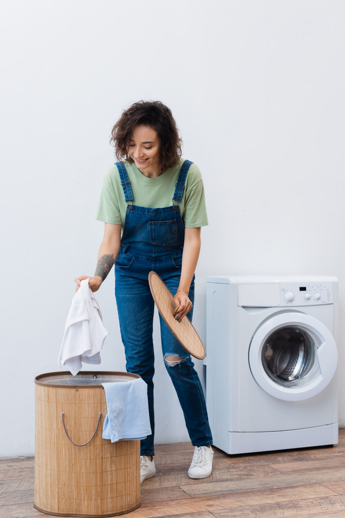 улыбающаяся женщина с прачечной возле корзины и стиральной машины - Фото, изображение