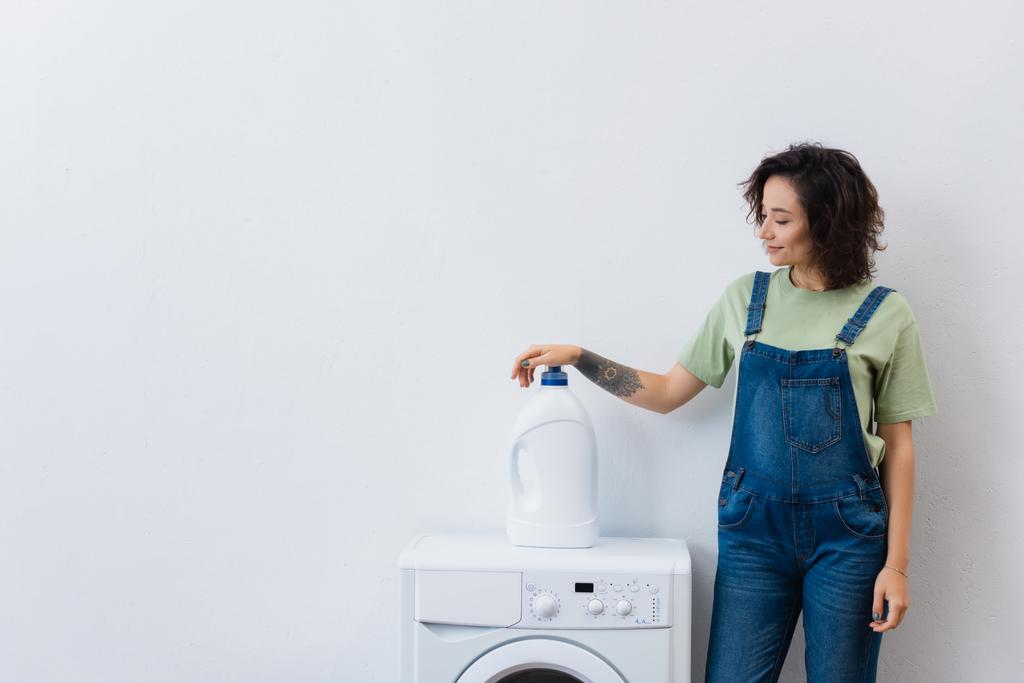 glimlachende huisvrouw staan in de buurt van fles wasmiddel op wasmachine - Foto, afbeelding