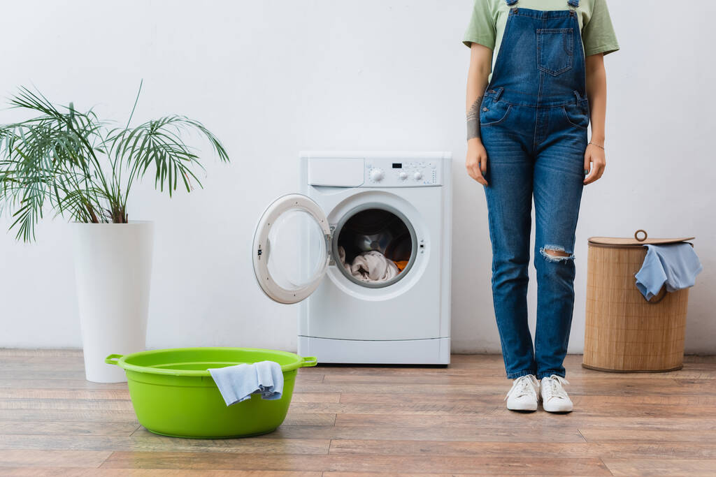 vista cortada da mulher em macacão jeans em pé perto da máquina de lavar roupa, bacia de roupa e cesta - Foto, Imagem