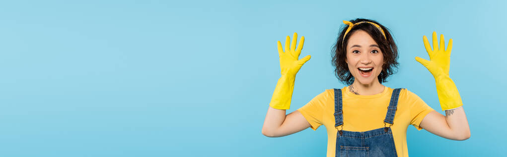 asombrado ama de casa mostrando las manos en guantes de goma amarilla aislados en azul, bandera - Foto, imagen
