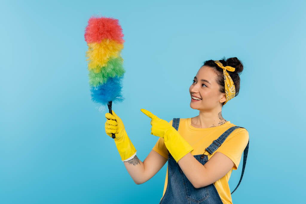 glimlachende huisvrouw in gele rubberen handschoenen wijzend naar kleurrijke stofborstel geïsoleerd op blauw - Foto, afbeelding