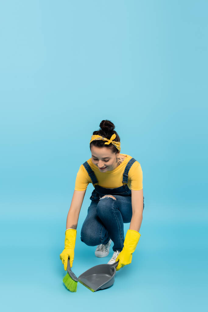 positive housewife in denim overalls and rubber gloves sweeping floor on blue - Photo, Image