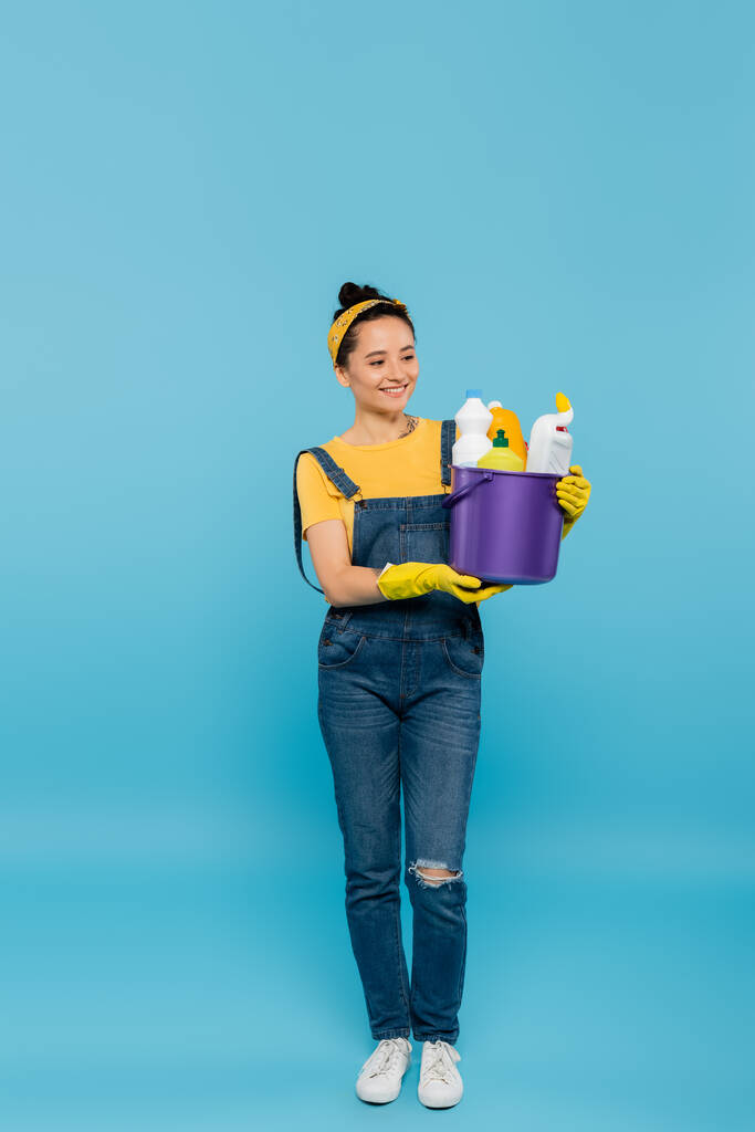 vista completa de ama de casa en overoles de mezclilla y guantes de goma amarillos que sostienen el cubo de detergentes en azul - Foto, Imagen