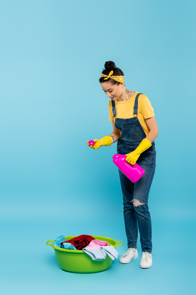 young housewife in denim overalls pouring detergent into laundry bowl on blue - Photo, Image