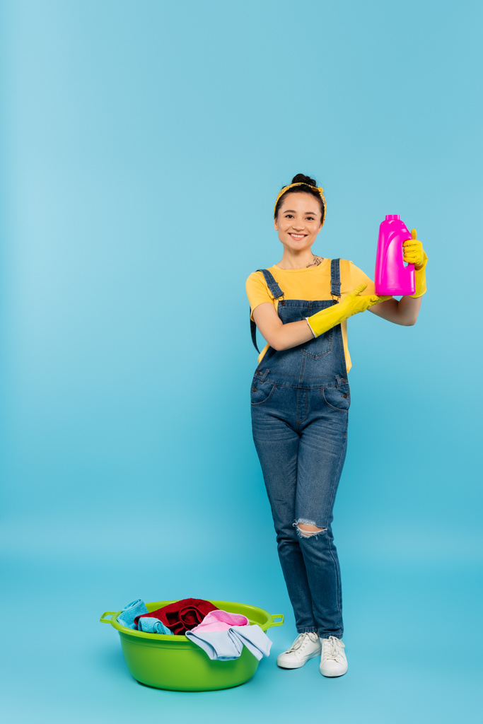 gelukkig huisvrouw in geel rubber handschoenen tonen fles wasmiddel in de buurt van waskom met kleren op blauw - Foto, afbeelding