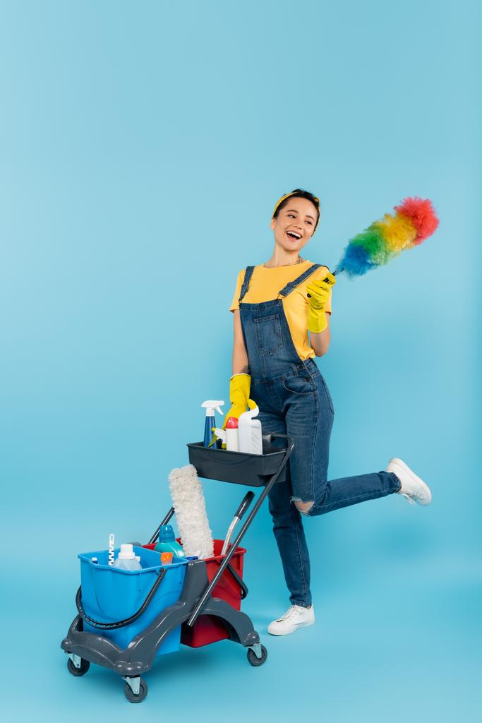 limpiador alegre con cepillo de polvo posando cerca del carro con detergentes y cubos en azul - Foto, imagen
