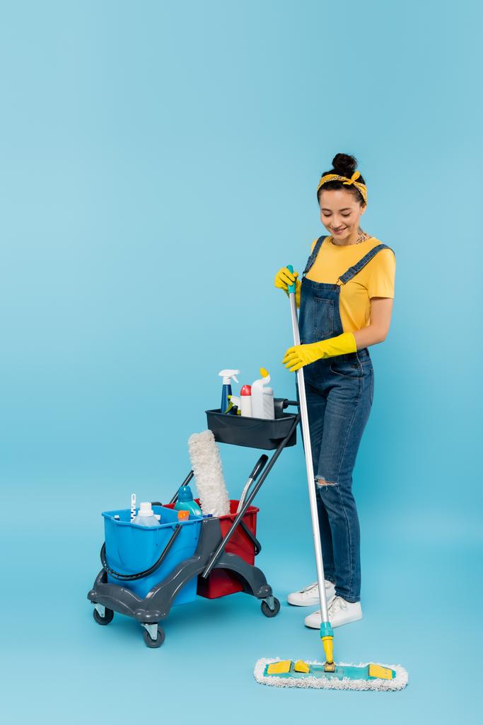 glimlachende reiniger met dweil nabij kar met emmers en wasmiddelen op blauw - Foto, afbeelding
