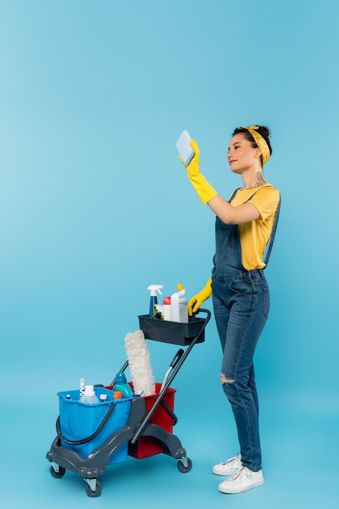 cleaner in denim overalls and rubber gloves taking selfie near cart with cleaning supplies on blue - Photo, Image