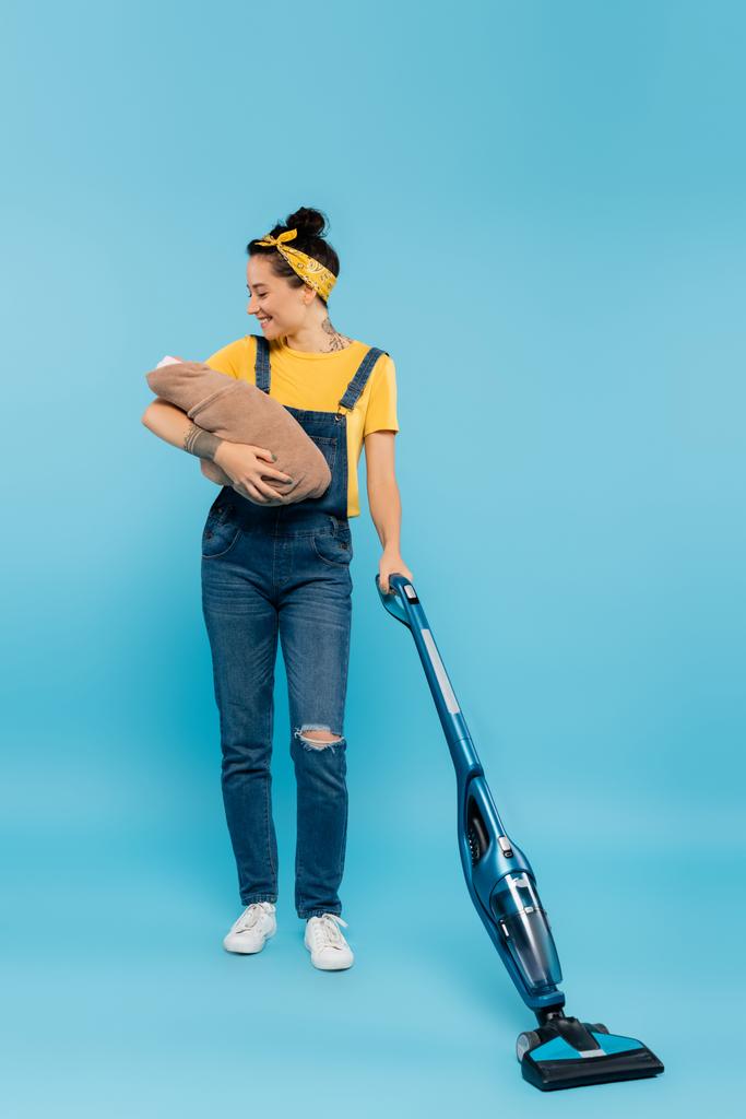 joyful woman in denim overalls holding baby doll while vacuuming on blue - Photo, Image