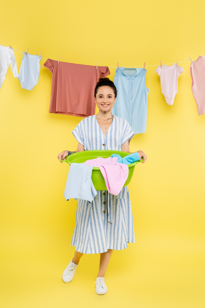 vista completa de ama de casa sonriente sosteniendo lavadero cerca de la ropa que cuelga en el fondo amarillo - Foto, imagen