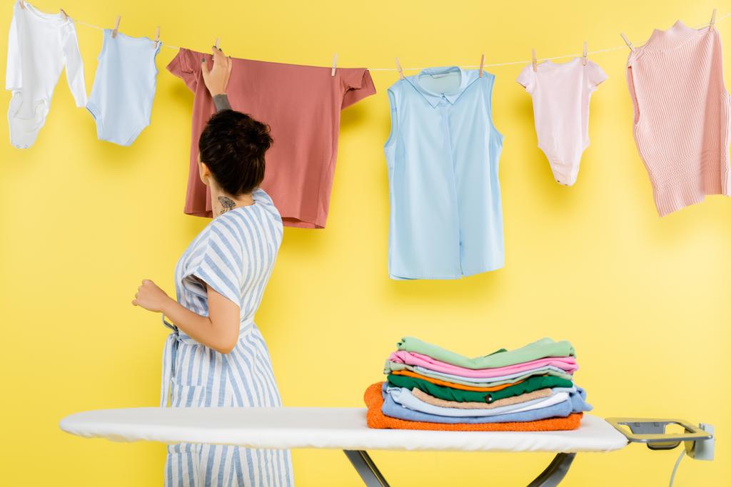 brunette woman touching hanging laundry near ironing board on yellow - Photo, Image