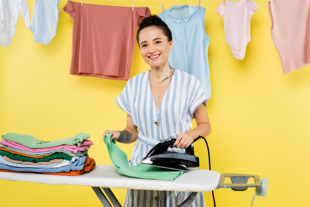 μελαχρινή γυναίκα χαμογελά στην κάμερα, ενώ σιδέρωμα ρούχα κοντά κρέμονται πλυντήριο σε κίτρινο - Φωτογραφία, εικόνα
