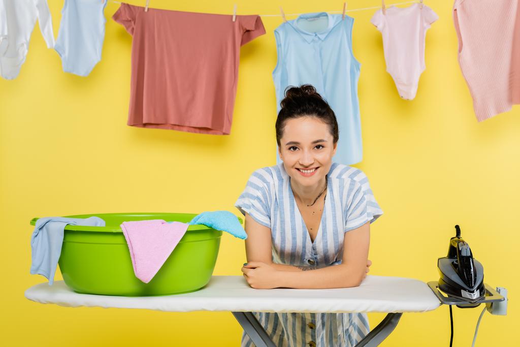 happy housewife looking at camera while leaning on ironing board near laundry bowl on yellow - Photo, Image