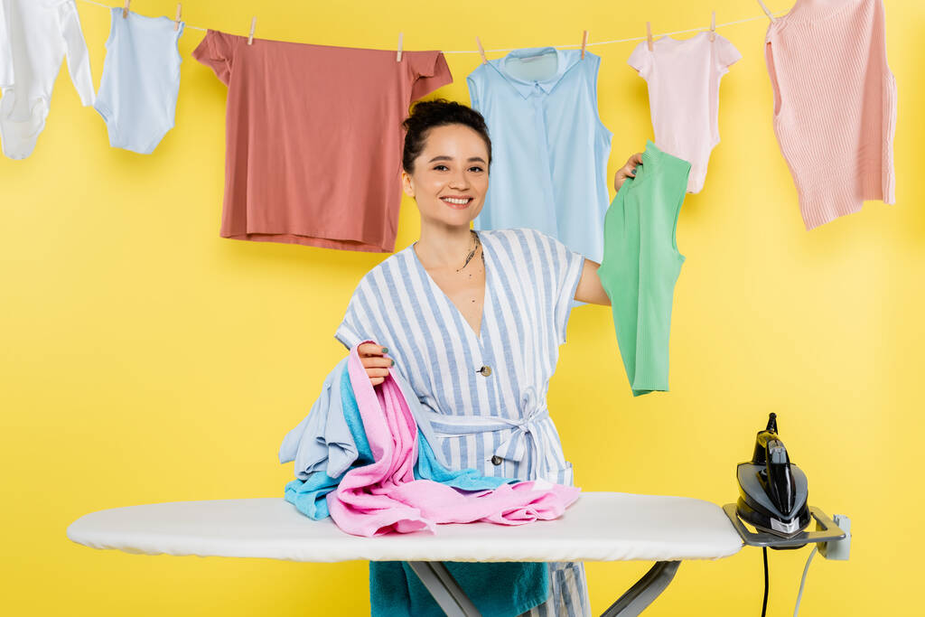 веселая домохозяйка смотрит в камеру, держа одежду возле гладильной доски на желтом - Фото, изображение