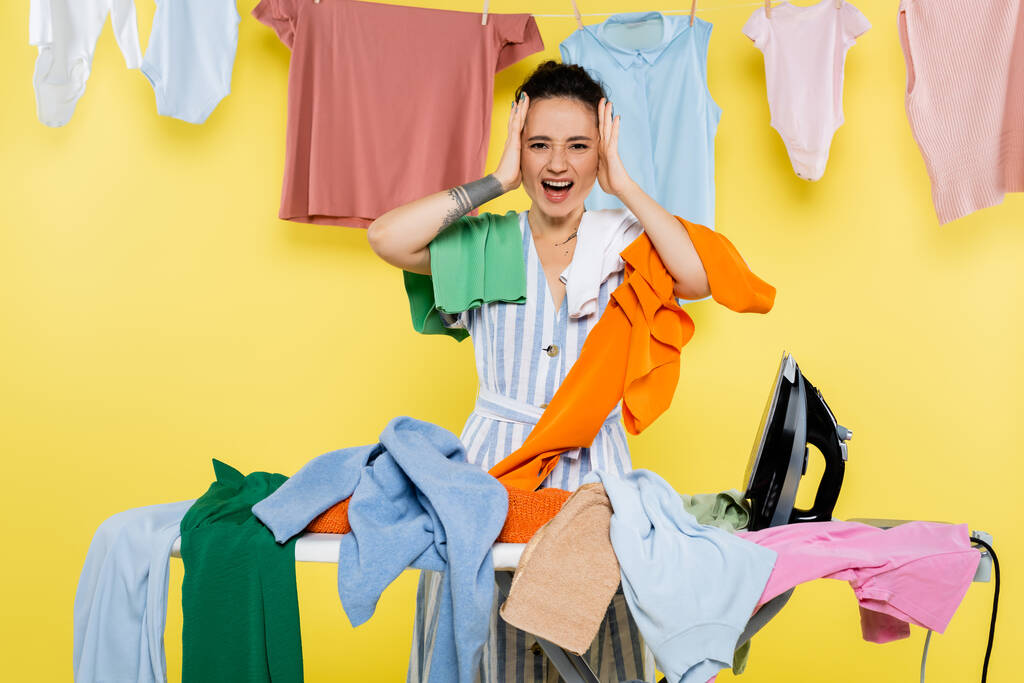 schockierte Hausfrau berührt Kopf beim Schreien in der Nähe von Wäsche auf Bügelbrett auf gelb - Foto, Bild