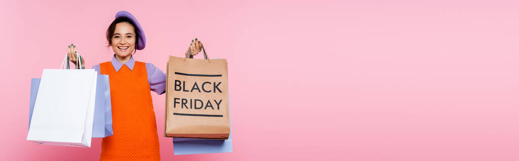 mujer sonriente sosteniendo compras y bolsa de compras con letras de viernes negro aislado en rosa, pancarta - Foto, imagen