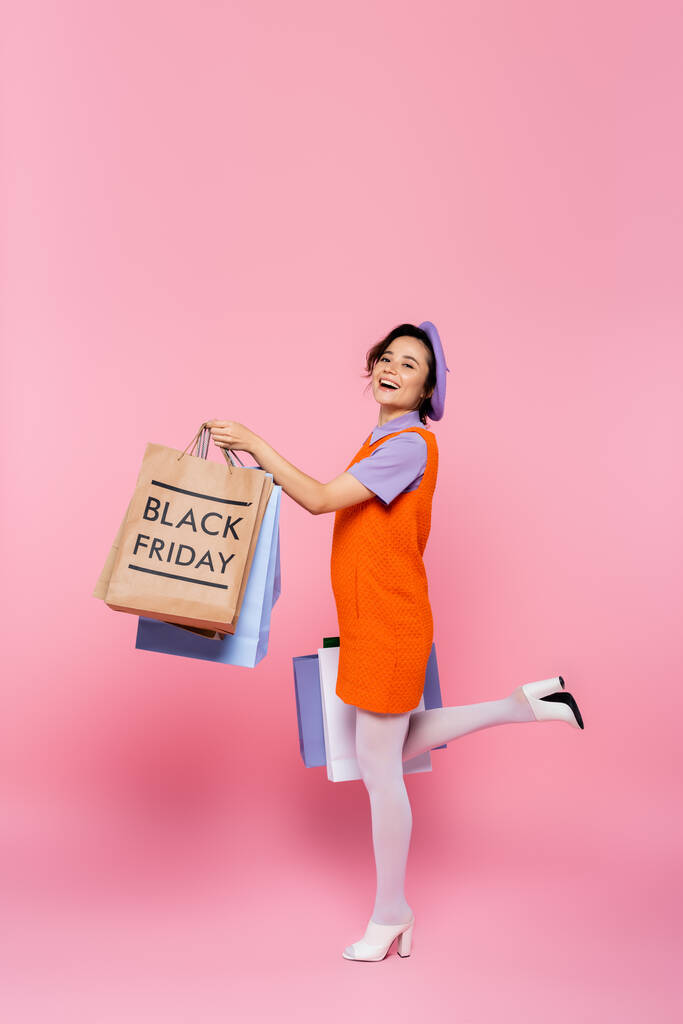 excited woman posing with purchases and shopping bag with black friday lettering on pink - Photo, Image