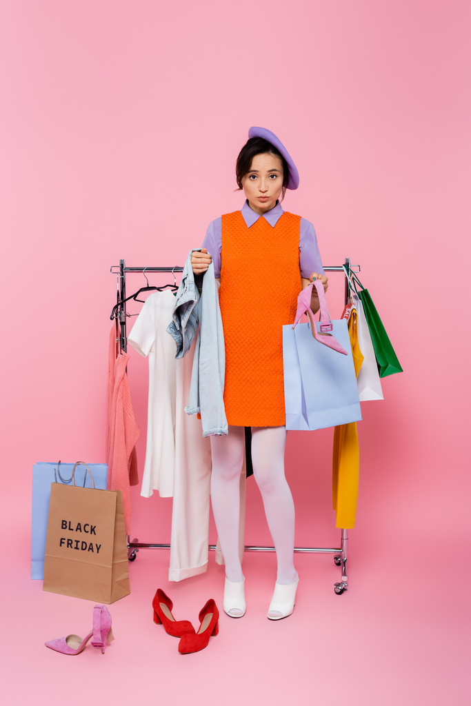 mujer confundida mirando la cámara cerca de rack con ropa y bolsas de compras en la venta de viernes negro en rosa - Foto, Imagen
