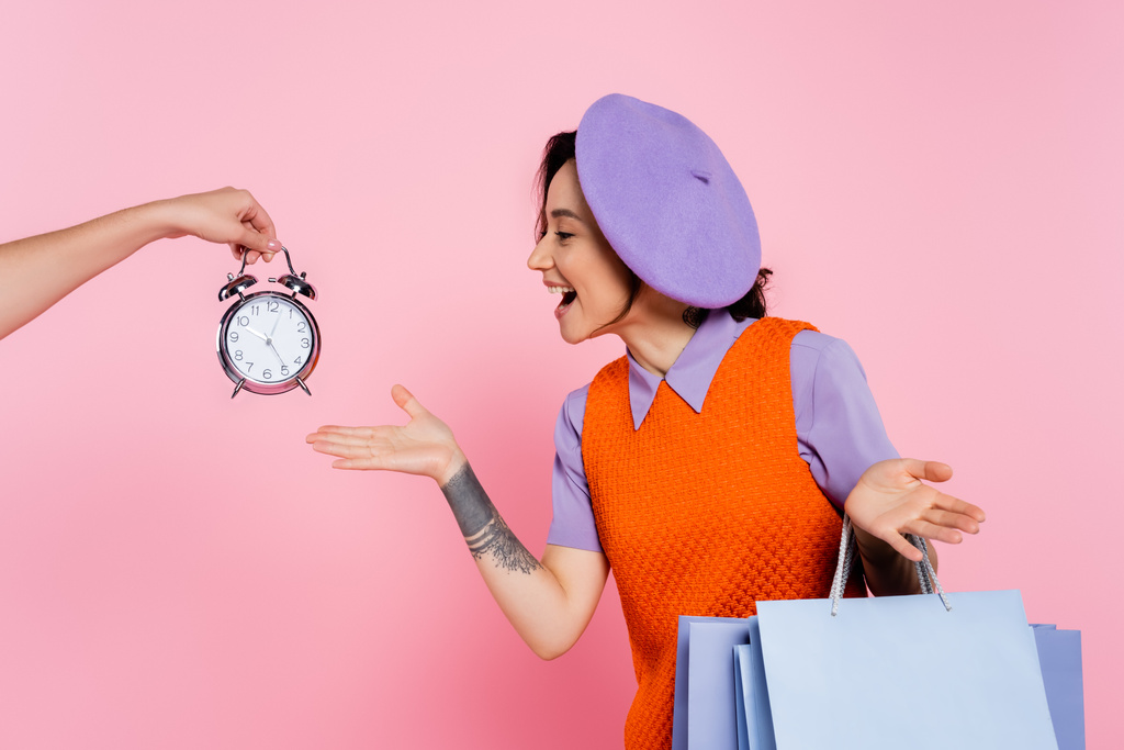 amazed woman showing wow gesture near female hand with vintage alarm clock isolated on pink - Photo, Image