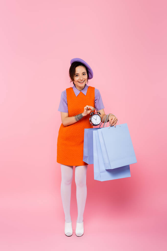 mujer feliz en vestido anaranjado sin mangas sosteniendo bolsas de compras y reloj despertador vintage en rosa - Foto, imagen