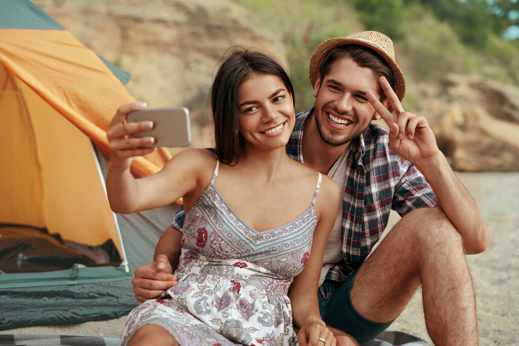 幸せな若いカップルはビーチで携帯電話で自撮りを取る。勝利の兆候を示す男。ロマンチックな関係の概念と一緒に時間を楽しむ。夏の自然の中でのキャンプ休暇 - 写真・画像