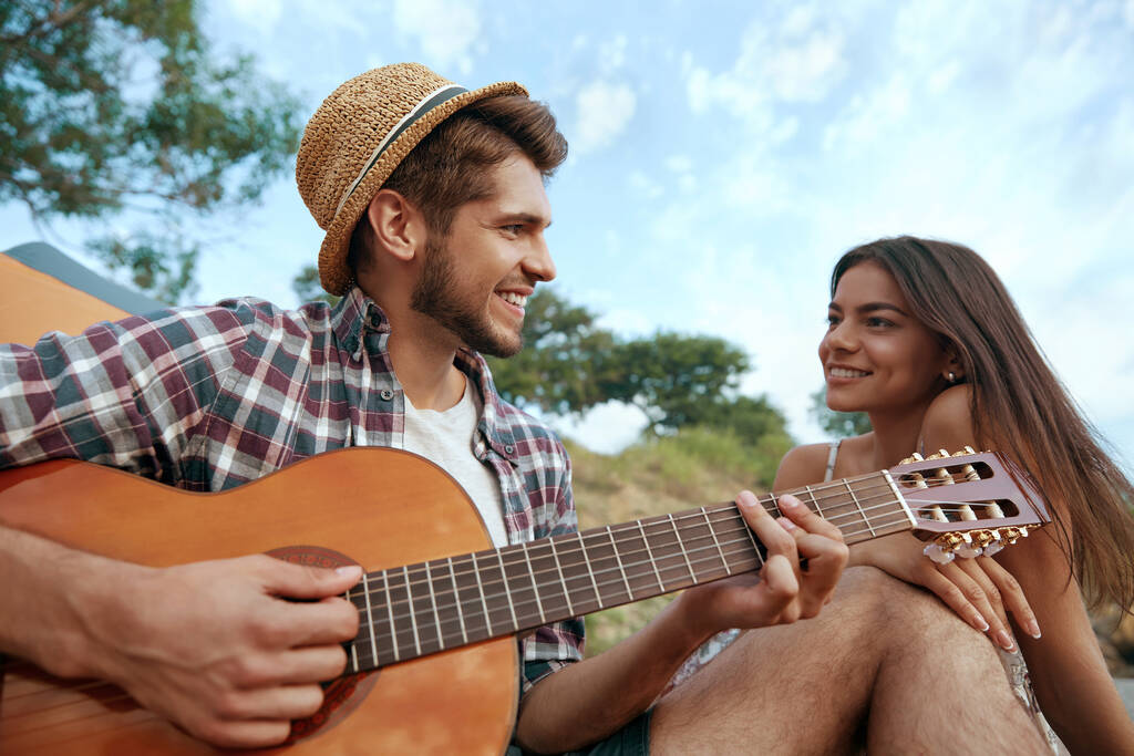 Uśmiechnięty facet grać na gitarze do pięknej zadowolonej dziewczyny. Młoda zakochana para Europejczyków odpoczywa na piaszczystej plaży. Pojęcie romantycznego związku i wspólnego spędzania czasu. Wakacje na łonie natury latem - Zdjęcie, obraz