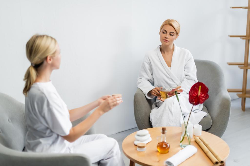 blonde client in bathrobe holding cup of tea while looking at blurred spa specialist with cream container - Photo, Image
