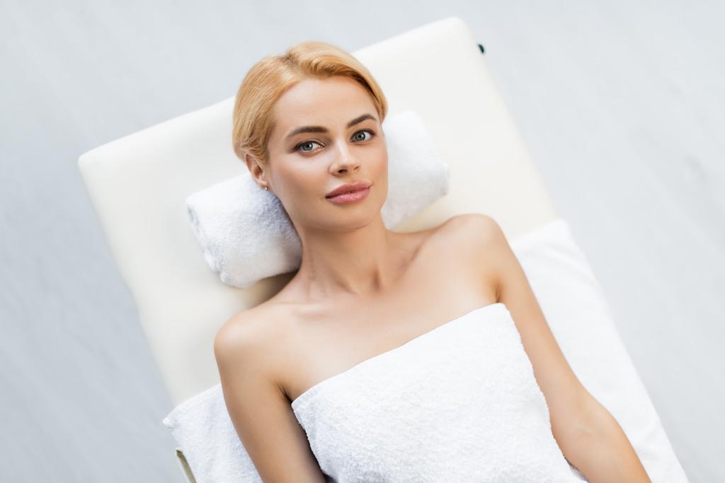 vista superior de la mujer rubia con los hombros desnudos acostado en la mesa de masaje en el centro de spa  - Foto, Imagen