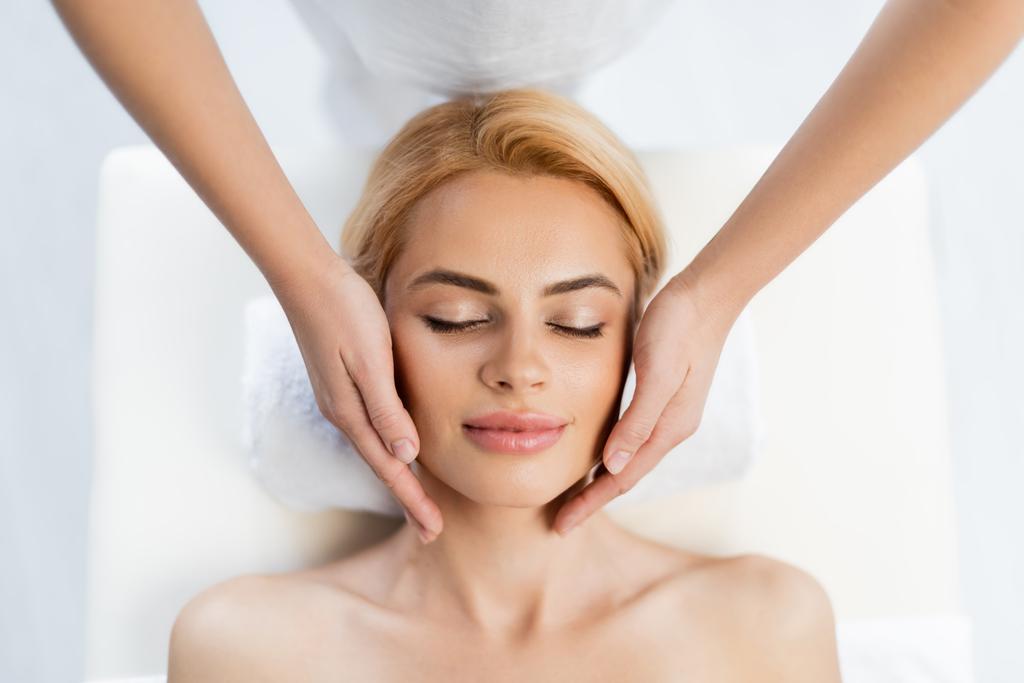 pohled shora na maséra při masáži obličeje blonďatého klienta  - Fotografie, Obrázek