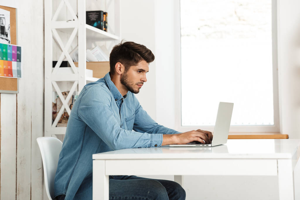 Giovane uomo bruna che lavora con il computer portatile mentre è seduto alla scrivania in ufficio al chiuso - Foto, immagini