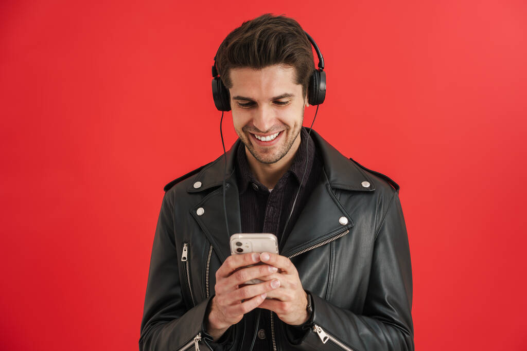 Молодий незахищений чоловік посміхається під час використання мобільних телефонів і навушників ізольовані над червоною стіною
 - Фото, зображення
